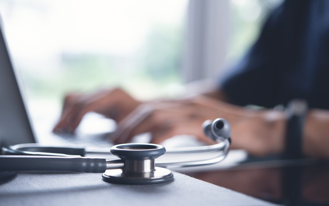 stethoscope on desk in foreground. medical professional in background working on computer